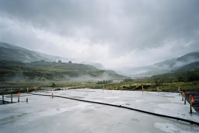 Illustrasjon: En stor, tom betongfundament i en norsk våt dal, merket med landmålerpinner, med én enkelt strømkabel som løper ut av bildet mot fjerne vannkraftturbiner under overcast himmel.