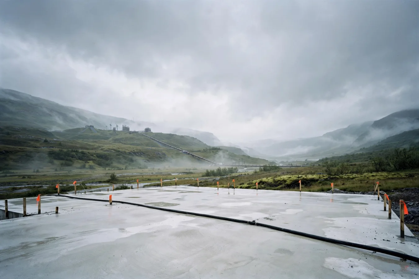 Illustration: A vast empty concrete foundation slab in a wet Norwegian valley, marked with survey stakes, a single power cable running out of frame toward distant hydroelectric turbines under an overcast sky.