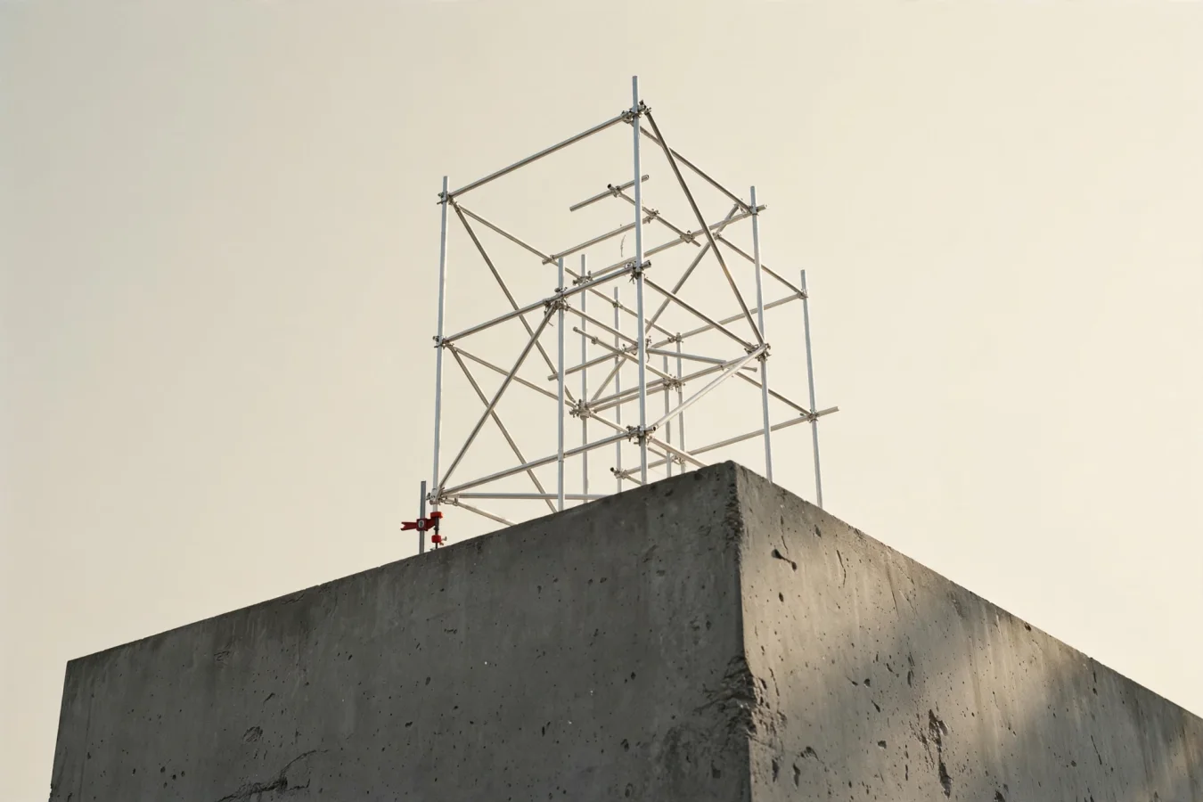 Skulpturell illustrasjon: et lite, intrikat aluminiumsskjelett bygget oppå en massiv betongfot, med én rød klemme i skjøten — illustrerer en modell bygget videre på andres basemodell.