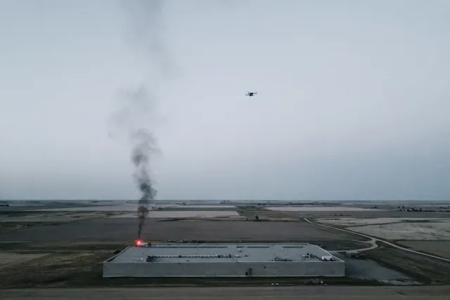 Illustration: a low data center building in flat rural terrain with dark smoke rising from the roof and a drone in the air above, in a muted editorial style.