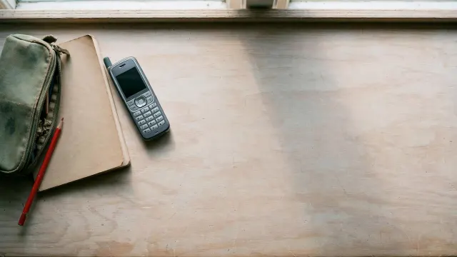 Illustrasjon: en skolepult i tre sett ovenfra, med blyantgraplappe, notatbok og en gammel telefon uten skjerm i bruk – resten av pulten er bevisst tom, som en forsvunnen enhet. Dæmpet nordisk dagslys, signalrød blyant som eneste fargekontrast.
