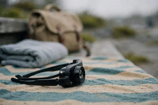 Illustrasjon: sorte smartbriller med kamera liggende på et håndkle på en offentlig strand, med treningstaske og skolesekk i bakgrunnen – et forbudssymbol i hverdagslige rom.