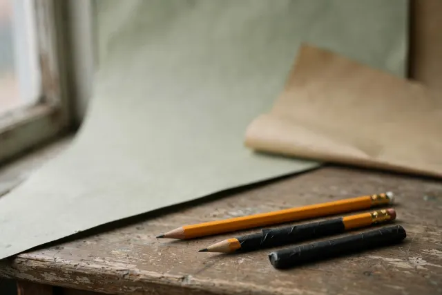 Illustrasjon: Tre blyanter på en skolepult etter lengde sortert, der den korteste er dekket av svart teip, den midterste delvis, og den lengste urørt – et bilde på aldersgrenser for AI i skolen.