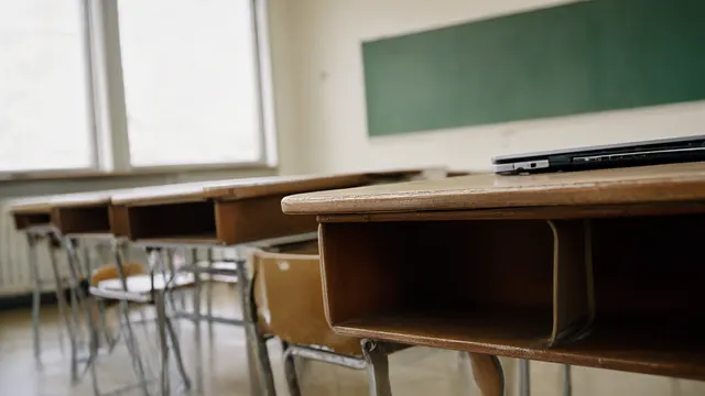 Illustrasjon: tomme skolebenker i et klasserom med dagslys; på én benk står en lukket laptop dyttet mot kanten, mens tavlen bak er utydelig.