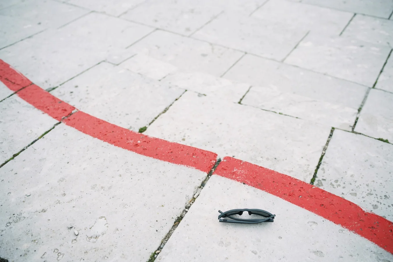 AI glasses lie face-down on stone pavement, cut off by a red barrier line curving across an empty public plaza – an illustration of a proposed temporary ban on AI glasses in public places.