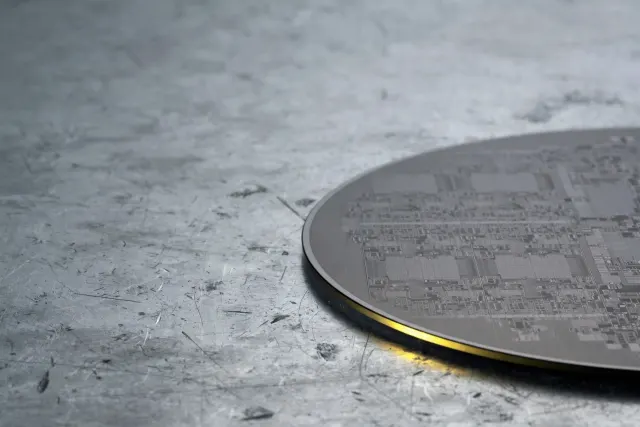 Illustration: a matte silicon wafer with fine circuit patterns rests on raw aluminum, lit by a narrow band of yellow light.