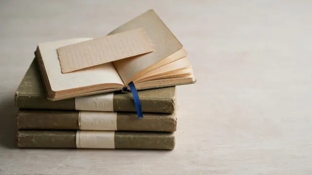 Stilleben av en stabel skolehefter der de nederste er forseglet med papirband, mens det øverste står halvåpent under en kartong som et slags oppsyn.