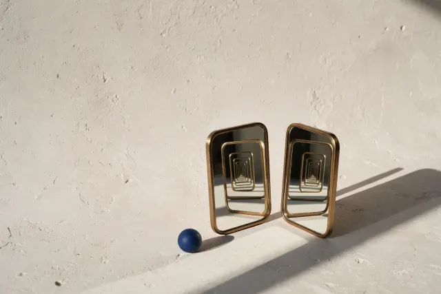 Two small mirrors facing each other on a cream surface creating an infinite reflection corridor, beside a small cobalt sphere. Illustration of a small language model modeling another's perspective.