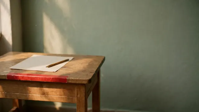 Illustrasjon: en liten skolepult i tre med blankt papir og blyant, avmerket med en rød tape som grense, i dempet dagslys.
