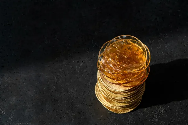 Illustration: a stack of gold-colored metal chips whose top half fades into translucent amber glass, a visual metaphor for funding pledges whose concrete value is partially unclear.
