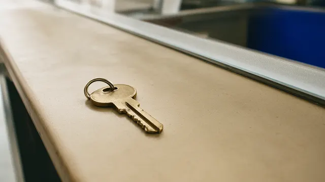 Illustration: A small brass checkout key rests on an empty shop counter, separated from the viewer by a panel of frosted glass – evoking AI agents that can now complete purchases, but only behind a confirmation barrier.
