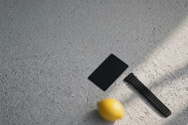 Two small black AI devices, a slim wristband and a credit-card-sized flat slab, resting on a gray surface in soft studio light.