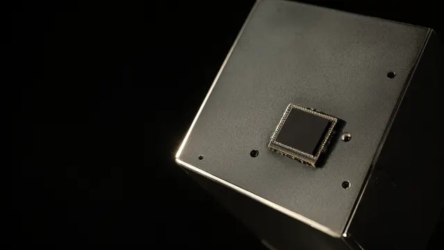 Illustrasjon: en mørk prosessorbrikke montert på en metallpanel fra en satellittprototyp, som fanger et hardt sollys mot sort rom.
