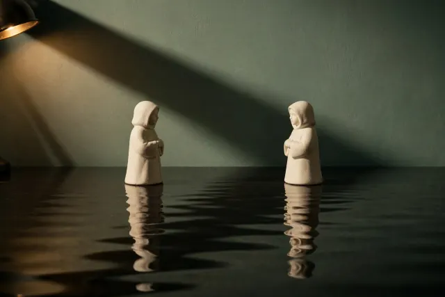 Illustration: two identical ceramic figurines facing each other on a dark reflective surface, the reflection subtly distorted, as a metaphor for companion chatbots amplifying a user's own thoughts.